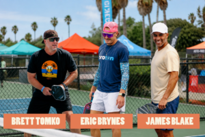 Three men with paddles posing on a fenced outdoor court during an event; orange name banners read Brett Tomko, Eric Brynes, James Blake, with tents in the background.