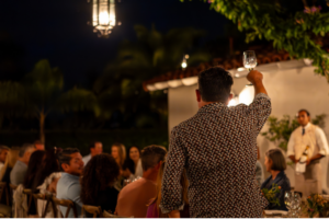 Man in patterned shirt raises a glass to toast at an outdoor nighttime dinner party with guests seated along a long table.